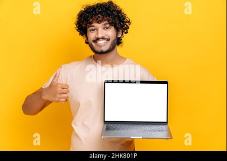 Joyeux Indien ou arabe gars dans un t-shirt de base, tient l'ordinateur portable ouvert avec écran blanc, montrant le pouce vers le haut, debout sur un arrière-plan isolé orange, souriant amical.Concept de maquette Banque D'Images