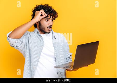 Excitée, perplexe, un homme indien ou arabe, dans une tenue décontractée, des lunettes à emporter, tient un ordinateur portable ouvert à la main, semble surpris et à l'écran avec la bouche ouverte, tout en se tenant sur un arrière-plan isolé orange Banque D'Images
