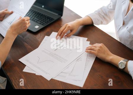 Vue rapprochée en grand angle des mains méconnaissables deux jeunes femmes d'affaires assises au bureau avec ordinateur portable et documents papier. Banque D'Images