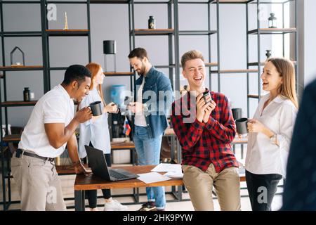 Une équipe de cinq jeunes collègues multiethniques et joyeux qui s'amusent à communiquer lors d'une réunion de groupe lors d'une pause-café dans une salle de bureau moderne. Banque D'Images
