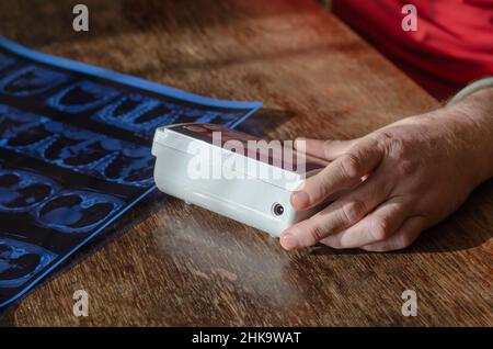 La main d'un homme tient un moniteur de tension artérielle numérique sur la table.Une acquisition CT des poumons se trouve à proximité.Mesure de la pression artérielle à la maison.Foyer sélectif Banque D'Images