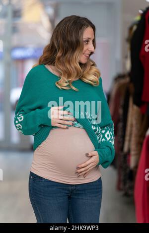 Jeune femme blonde enceinte essayant de porter des vêtements dans un magasin de vêtements Banque D'Images