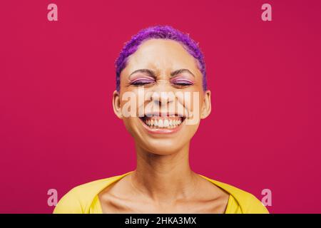 Vibes vibrantes.Jeune femme excitée souriant avec ses yeux fermés tout en se tenant contre un fond rose.Jeune femme tendance portant un maquillage avec Banque D'Images