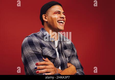 Un jeune homme insouciant souriant avec ses yeux fermés dans un studio.Un jeune homme heureux debout seul sur fond rouge.Jeune homme tendance portant une na Banque D'Images