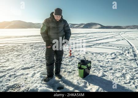 Un pêcheur pêche dans un trou sur la glace d'un grand lac gelé par une journée ensoleillée.La joie de la pêche d'hiver sur le lac Baikal Banque D'Images