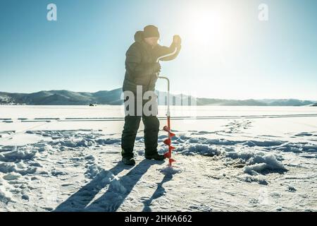 Un pêcheur fore un trou dans la glace d'un grand lac gelé par une journée ensoleillée.La joie de la pêche d'hiver Banque D'Images