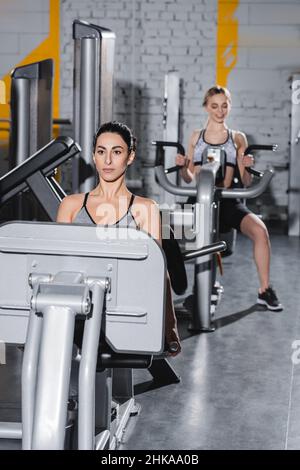 Jeune sportif du Moyen-Orient travaillant sur la machine de presse de jambe près d'un ami flou dans la salle de sport Banque D'Images