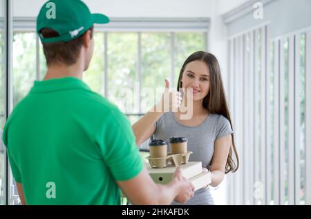 Le personnel de livraison en uniforme vert a remis la boîte à pizza et la tasse de café au jeune client.La femme a levé ses pouces satisfaits de la nourriture d Banque D'Images