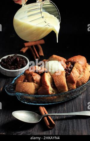 Versez la vanille VLA dans le pudding de pain frais fait de baguette en tranches avec raisin et cannelle, sur la table en bois noir Banque D'Images