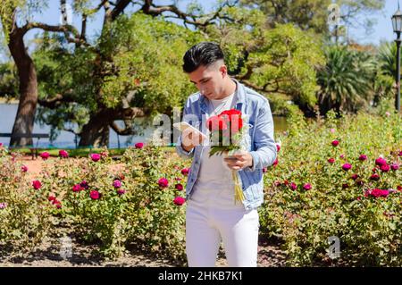 homme caucasien debout dans le jardin d'un parc avec beaucoup de fleurs et d'arbres derrière lui, avec un bouquet de fleurs dans sa main s'inquiétait de lire des messages Banque D'Images