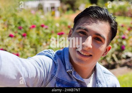 portrait en gros plan d'un jeune homme hispanique de race blanche latino-argentin prenant un selfie à l'extérieur, dans un parc public avec un jardin avec des roses sur le backgroun Banque D'Images