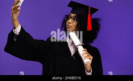 jeune étudiant afro-américain en lunettes et en chapeau de graduation prenant le selfie isolé sur le violet Banque D'Images