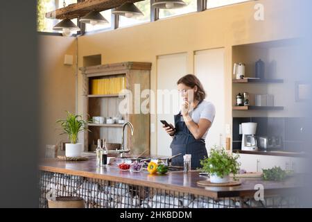 Femme enceinte caucasienne portant un tablier en utilisant un smartphone dans la cuisine à la maison Banque D'Images