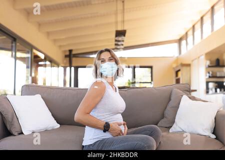 Portrait d'une femme enceinte caucasienne portant un masque facial touchant son ventre assis sur le canapé Banque D'Images