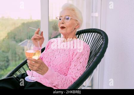 Ancienne femme élégante sénior sur la terrasse fumeur cigarette avec un verre de vin blanc.Mauvaise habitude, concept de dépendance Banque D'Images