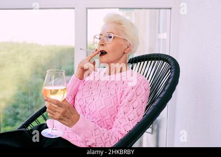 Ancienne femme élégante sénior sur la terrasse fumeur cigarette avec un verre de vin blanc.Mauvaise habitude, concept de dépendance Banque D'Images