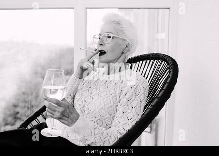 Ancienne femme élégante sénior sur la terrasse fumeur cigarette avec un verre de vin blanc.Mauvaise habitude, concept de dépendance Banque D'Images