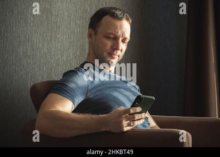 Portrait d'un jeune adulte européen sérieux assis avec un smartphone dans un canapé devant la fenêtre le matin ensoleillé Banque D'Images