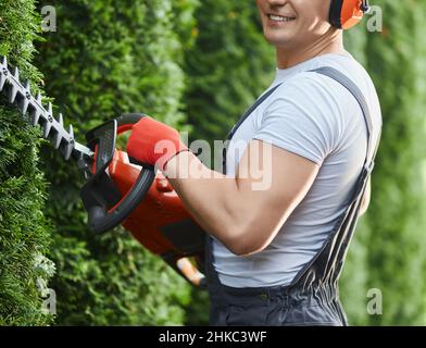 Gros plan d'un jardinier musclé dans une combinaison et des gants à l'aide d'une machine à tondre électrique.Homme caucasien paysagant et prenant soin des plantes à l'extérieur. Banque D'Images