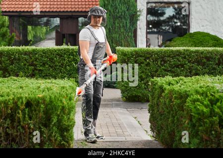 Jardinier professionnel de sexe masculin utilisant une machine à découper électrique pour le travail dans le jardin.Homme caucasien portant une combinaison brune, des gants et des lunettes de protection. Banque D'Images