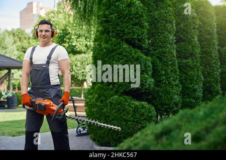 Jardinier professionnel de sexe masculin portant une combinaison, des gants et des lunettes bruns debout dans un jardin d'été avec taille-haie électrique.Homme qui apprécie le travail saisonnier dans la cour arrière. Banque D'Images