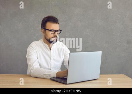 Jeune homme d'affaires travaillant sur un ordinateur portable assis au bureau sur fond gris Banque D'Images