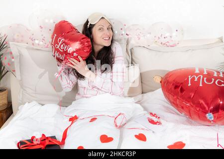 Une belle femme en pyjama rose avec un ballon en forme de coeur se trouve sur le lit à la maison.Femme le jour de la Saint-Valentin, anniversaire ou anniversaire.Symbole d'amour Banque D'Images