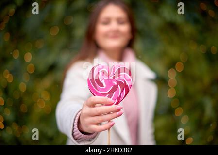 bonbons au caramel en forme de cœur dans la main de fille.Flou belle femme prolongeant rouge coeur-forme loolly-pop sur bâton à l'appareil photo Banque D'Images