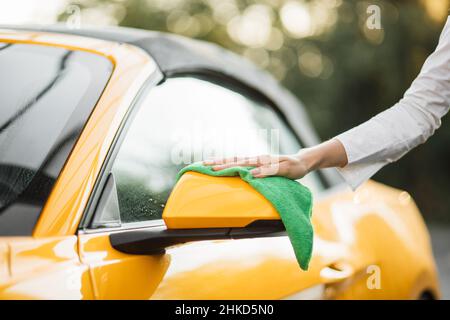 Gros plan de la main d'une jeune femme caucasienne essuyant le rétroviseur intérieur de sa voiture de luxe moderne dans une station de lavage de voiture en libre-service à l'extérieur avec un chiffon en microfibre verte en été matin ensoleillé Banque D'Images