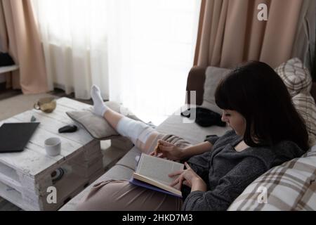 Jeune femme après une opération ACL, assise sur le canapé et lisant un livre. Banque D'Images