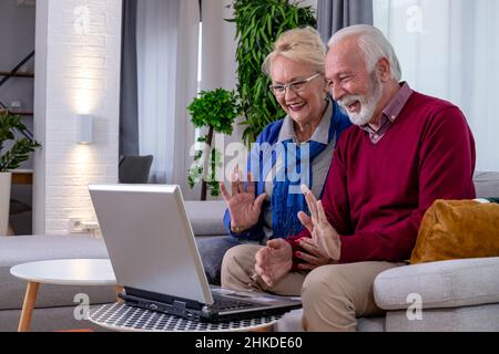 Un charmant couple senior est assis sur un canapé et parle à leurs enfants et petits-enfants par appel vidéo sur un ordinateur portable dans leur salon. Banque D'Images