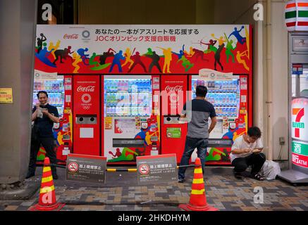 tokyo, japon - août 30 2021 : des hommes japonais mangeant des fast-foods de konbini devant un distributeur de boissons enveloppé de vecteurs représentant des sports sil Banque D'Images