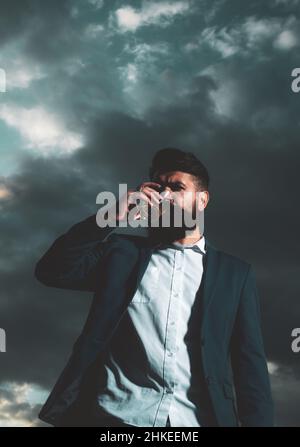 Un homme à la barbe et à la moustache tient une boisson alcoolisée sur fond de ciel. Service et alcoolisme concept. Banque D'Images