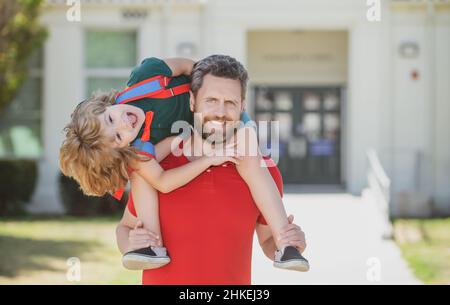 Professeur en t-shirt et adorable écolier avec sac à dos près du parc scolaire.L'homme et l'excitée Kid Kid piggyback émerveillé près de l'école. Banque D'Images