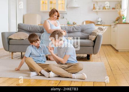 Les enfants deux frères jouent ensemble sur le sol, tandis que la jeune mère s'assoit à la maison sur le canapé dans la salle de séjour contre la cuisine de fond, les garçons ont l'amitié amusante entre les frères loisirs en famille dans la salle de séjour Banque D'Images