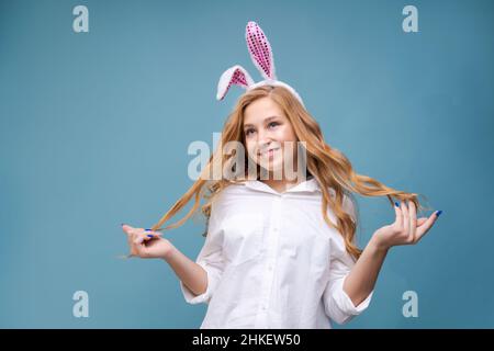 Jeune belle femme dans les oreilles de lapin de pâques mignons fait un geste normal, joue avec sa main avec de longs cheveux, dans une chemise blanche sur un fond bleu sourit heureux.Fille caucasienne dans le studio Banque D'Images