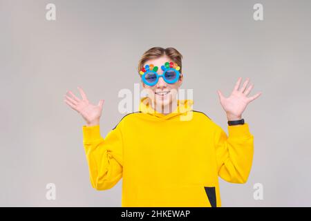 Photo gai excitée jeune gars vêtu d'un sweat-shirt jaune et de lunettes bleues avec l'inscription anniversaire, sourit et fait différents gestes avec ses mains sur les côtés sur un fond gris Banque D'Images