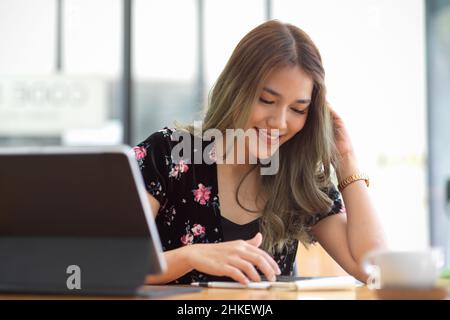 Jolie charmante jeune femme asiatique assise devant son espace de travail et utilisant un smartphone.Femme d'affaires se détend sur l'application mobile. Banque D'Images