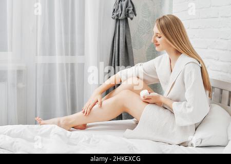 Jolie femme met de la crème après-rasage sur sa jambe assise sur le lit en profil Banque D'Images