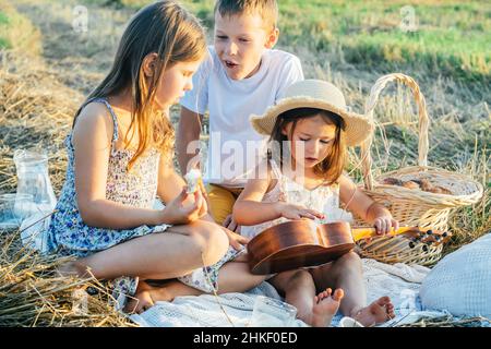 Portrait d'un garçon joyeux et de deux filles assis sur une couverture dans le champ, ayant pique-nique, mangeant du pain.Un moment de détente.Jouer de la guitare, apprendre à jouer ukulele Banque D'Images