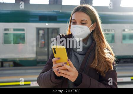 Jeune femme avec masque médical acheter un billet en ligne avec smartphone à la gare Banque D'Images