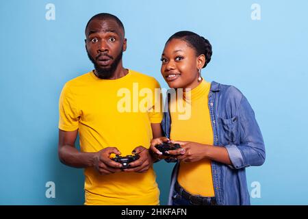 Homme et femme utilisant jpystick pour jouer à des jeux vidéo en studio, ayant le plaisir dans la relation.Heureux couple jouant en ligne jeu avec contrôleur sur la console, debout sur fond bleu. Banque D'Images