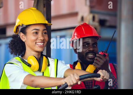 Deux hommes et femmes afro-américains travaillant en uniforme et avec un casque, sur un chariot élévateur à conteneur diesel, sur un quai commercial. Banque D'Images