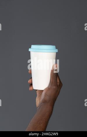 Main d'homme africain avec une tasse en carton ou une tasse jetable pour le café ou le thé.Gobelet blanc jetable avec fil en plastique bleu dans la main de l'homme.Concept de boîtes jetables.Copier l'espace. Banque D'Images