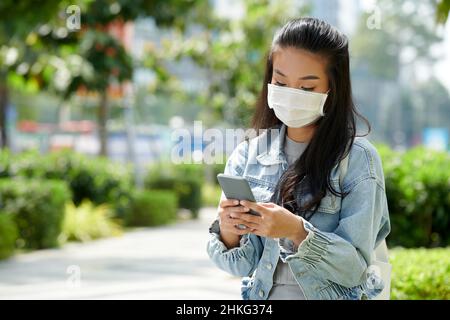 Jeune femme en masque de protection assise dans le parc et amis à envoyer des SMS Banque D'Images
