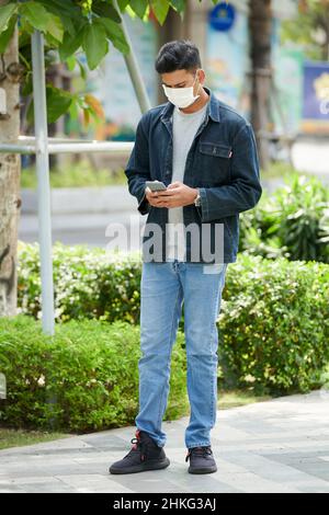 Jeune homme dans le masque de protection debout dans le parc et la lecture de messages texte de la petite amie qui est en retard pour une date Banque D'Images
