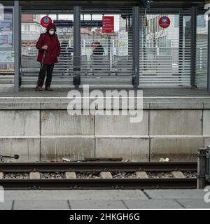 Une dame portant un masque Covid est debout avec deux bâtons de marche dans une salle d'attente sur une plate-forme.Au-dessus de sa tête le signe d'avertissement: Distance. Banque D'Images