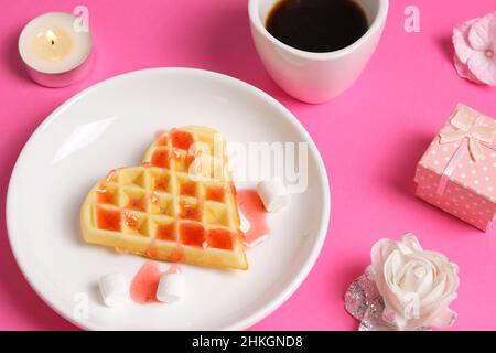 Vue rapprochée sur les gaufres faites maison en forme de cœur recouvertes de sirop de fraise, une tasse de café et une boîte cadeau.Petit déjeuner romantique pour la Saint-Valentin Banque D'Images
