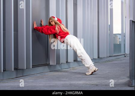 Une femme afro-américaine joyeuse dans des vêtements de sport rouges et portant des écouteurs poussant un mur en métal gris Banque D'Images