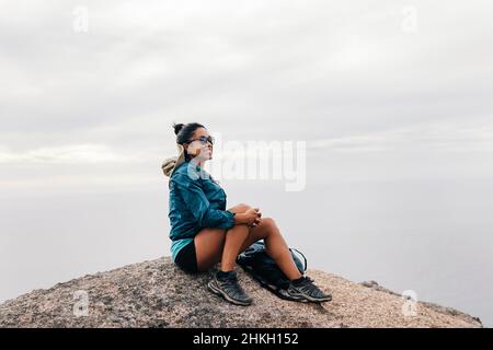 Femme souriante assise sur la falaise et regardant la vue.Une rancheuse féminine se reposant sur un rocher qui s'éloigne. Banque D'Images